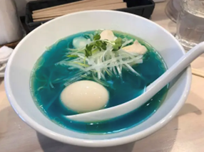 a white bowl and spoon with noodles, greens, and an egg in a blue broth